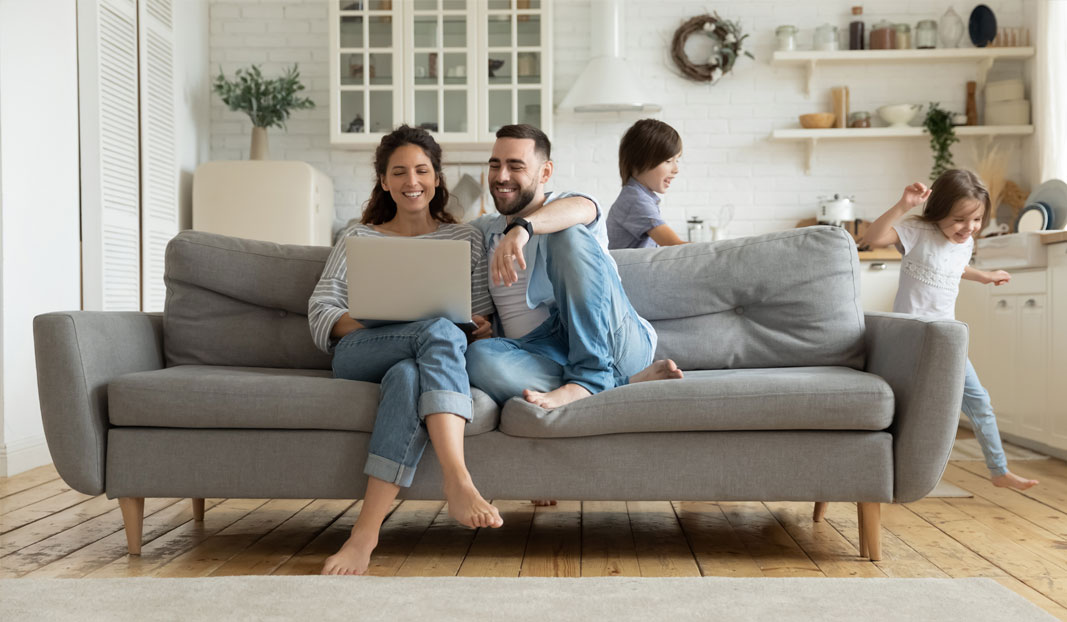 Mum and dad sitting on a sofa using a laptop while children play 